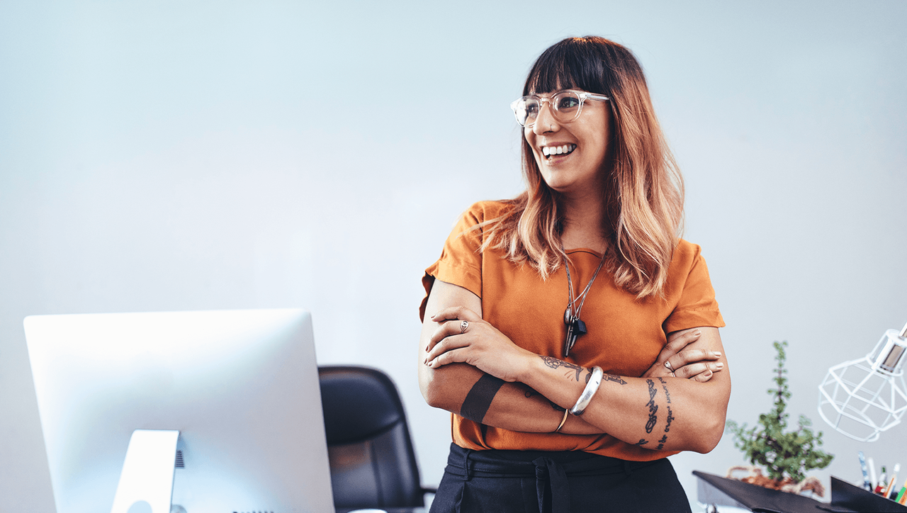 Blonde woman in gold shirt stands behind a computer in an office, arms folder at her chest, smiling and looking to the side