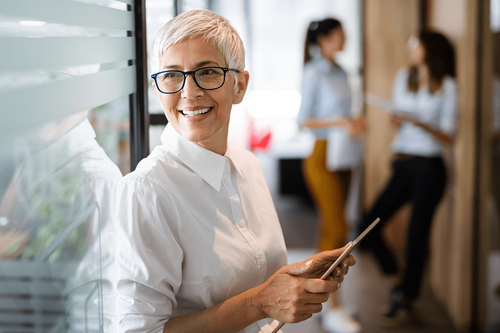 A white older woman with dark rimmed glass and a white button up shirt holds a tablet in her hands. She stands next to a glass office door and gazes to her side smiling.