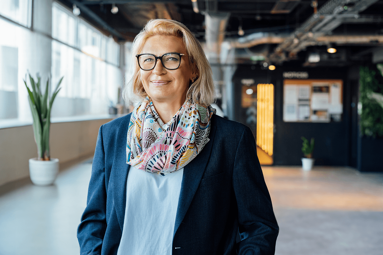 A white middle aged woman stands smiling slightly directly at the camera. She stands in an office setting with windows behind her. She wears a patterned scarf, a white shirt and a dark blue blazer with her hands in her pockets.