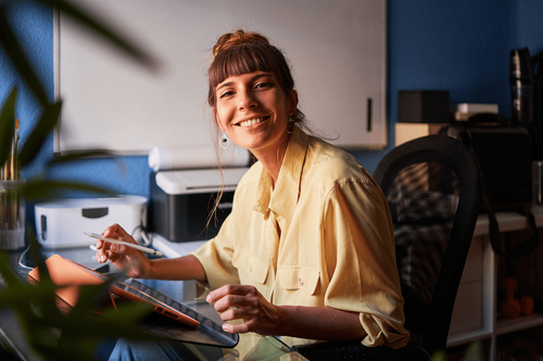 A young female leader smiles, pivoted to her right toward the camera. She's sitting at an office desk. Her hair is up in a neat bun and she's wearing a warm yellow button up shirt with the sleeves rolled up.