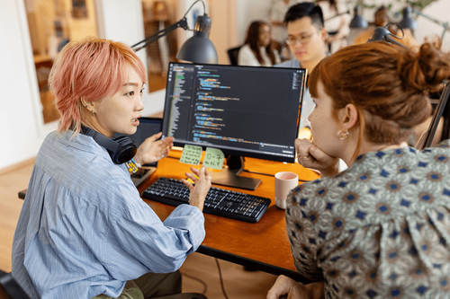 Two people interact facing a desktop monitor in an office setting.