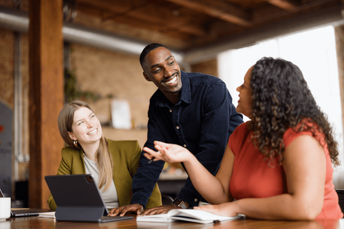 Three professionals smile and collaborate around a table with a tablet and notebook.